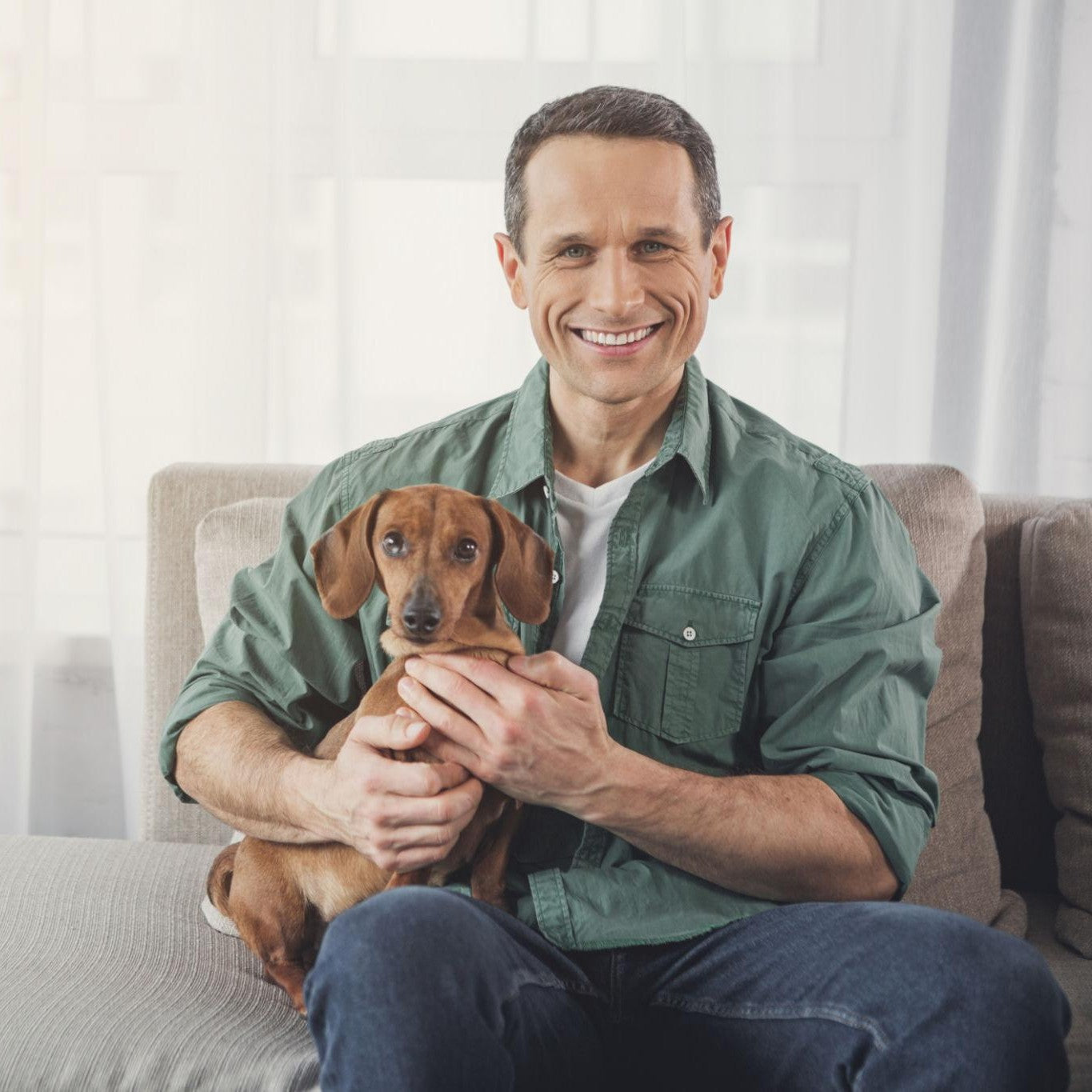 Man sitting on a couch holding a small brown dog, smiling at the camera.